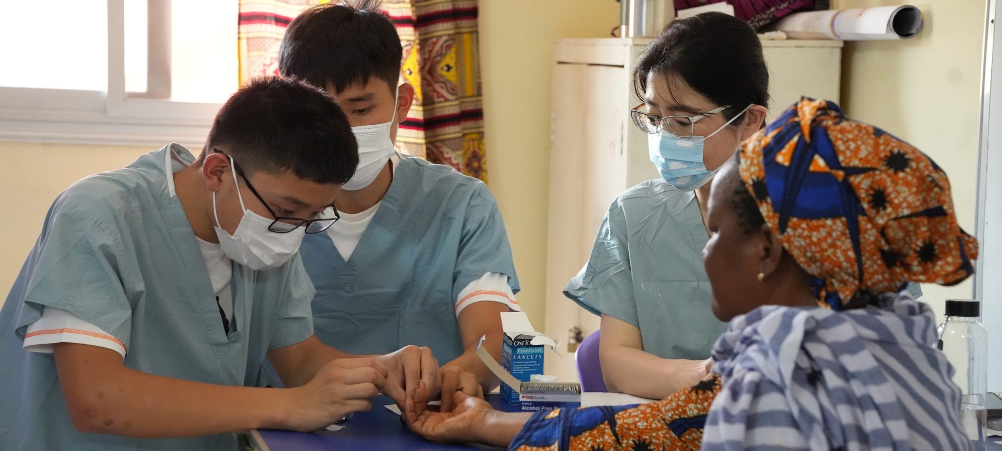 Three ACO clinicians in scrubs testing a patient's blood sugar at a village clinic table in Senegal