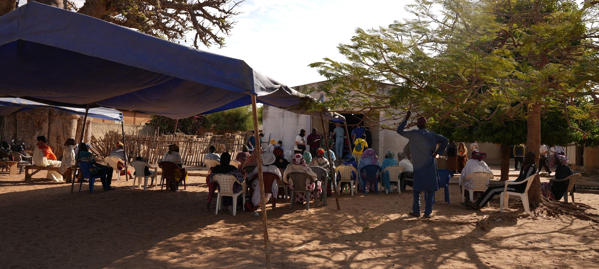Patients waiting on plastic chairs under a blue tarpaulin at an open-air ACO clinic in Senegal, with the clinic building behind them