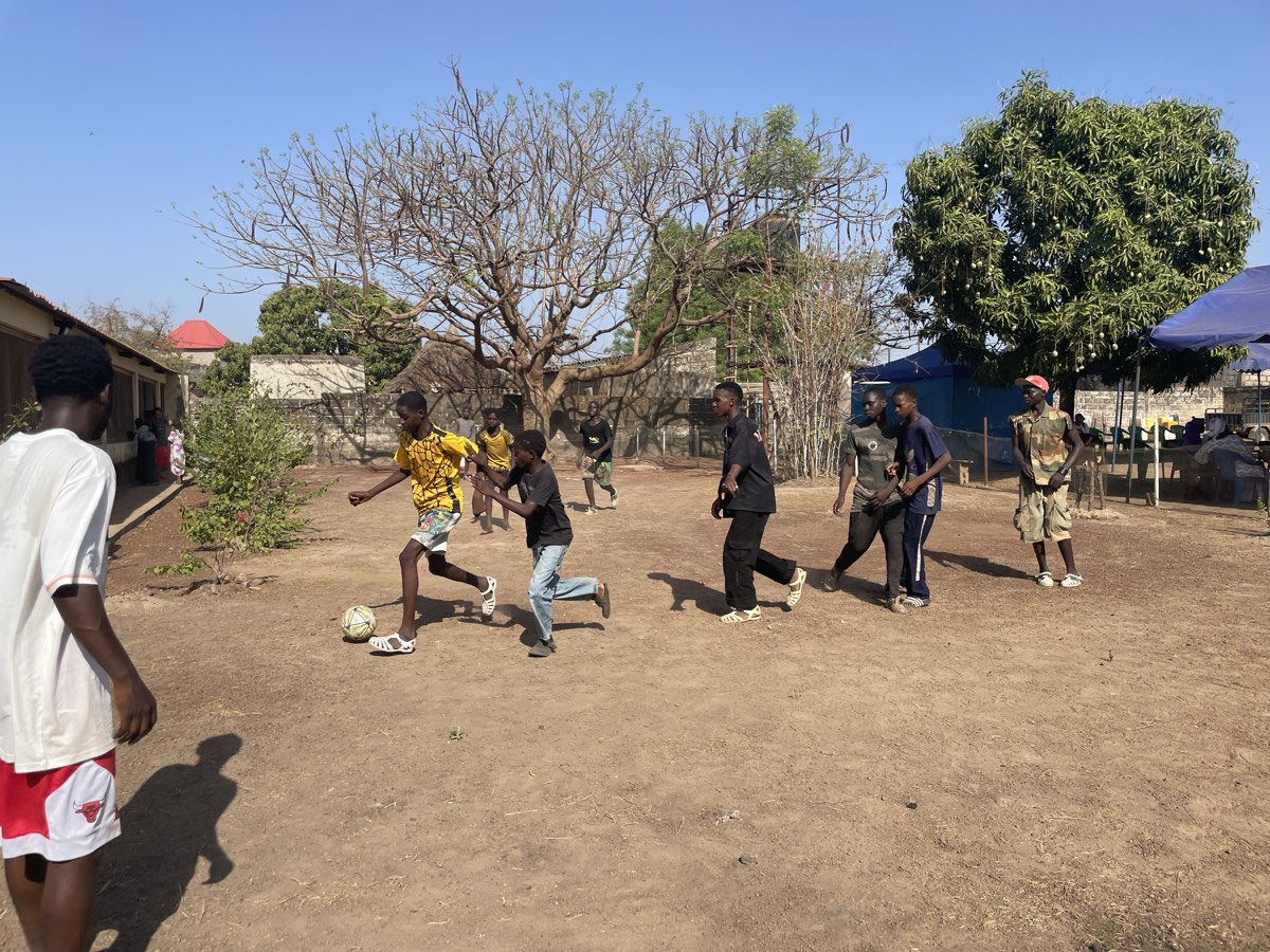 Boys sprinting for the ball on a bare earth pitch under trees at Fadiga