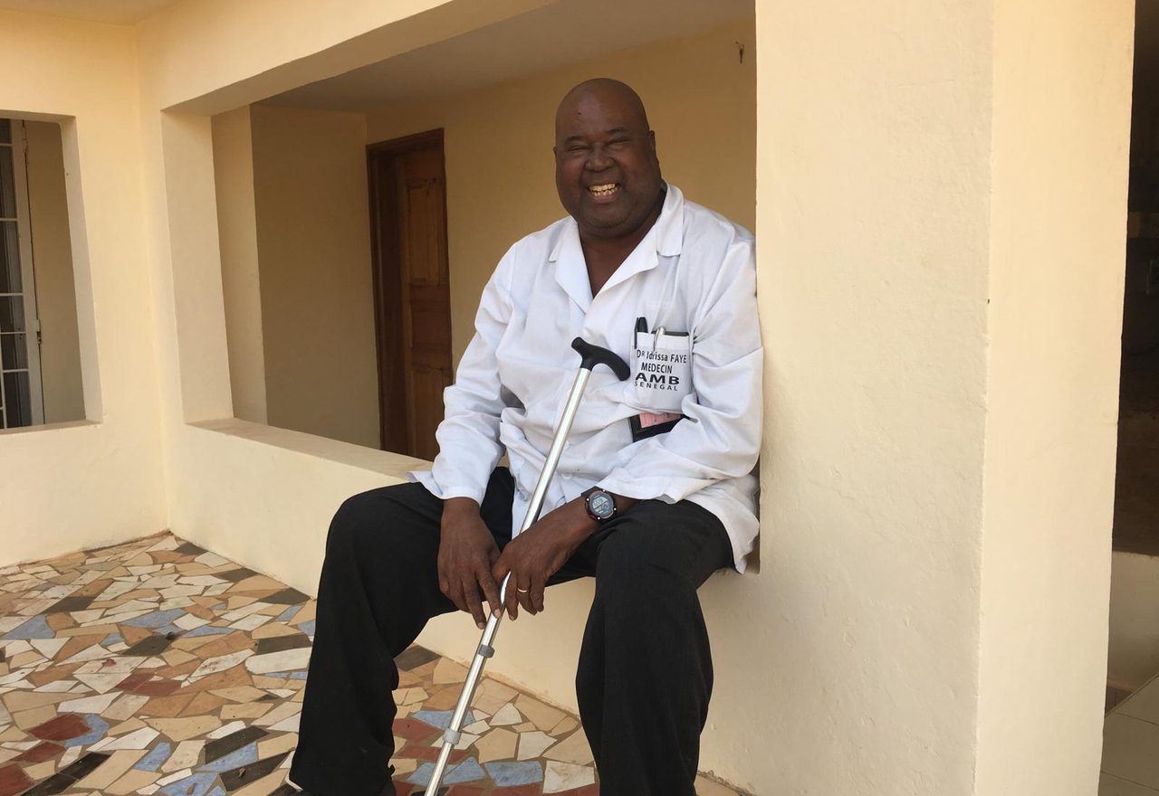 Dr. Idrissa Faye seated outside the clinic in his white coat, smiling, his walking stick in his hands.