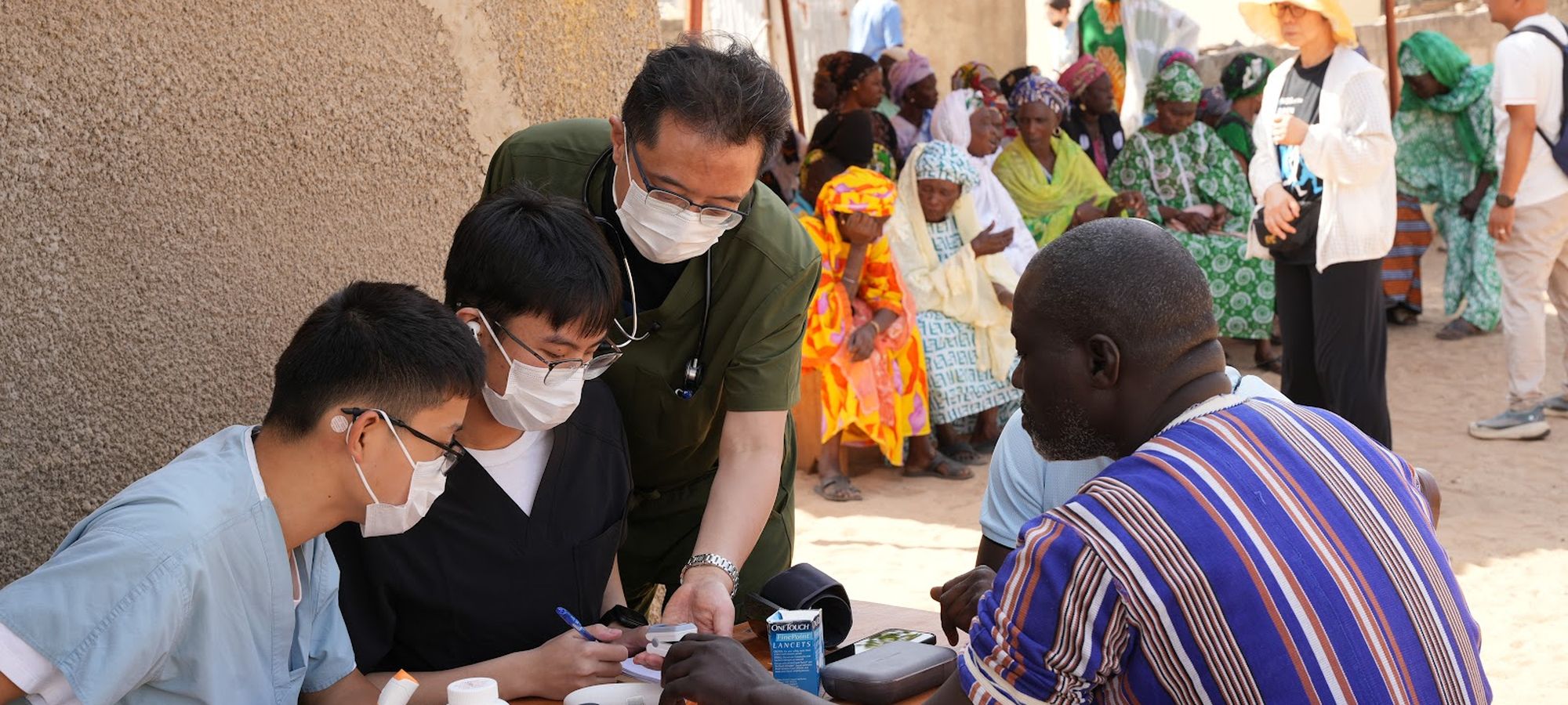 ACO clinicians examining a patient at a village outreach clinic in Senegal, with community members waiting behind them