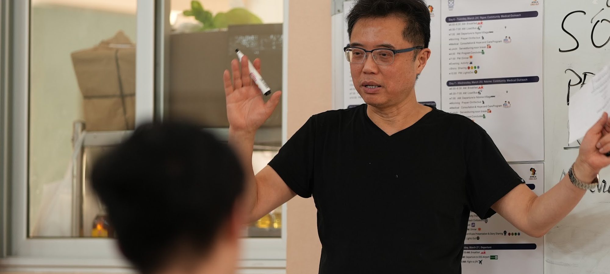 An ACO physician teaching at a whiteboard while youth volunteers look on, the trip schedule posted beside him
