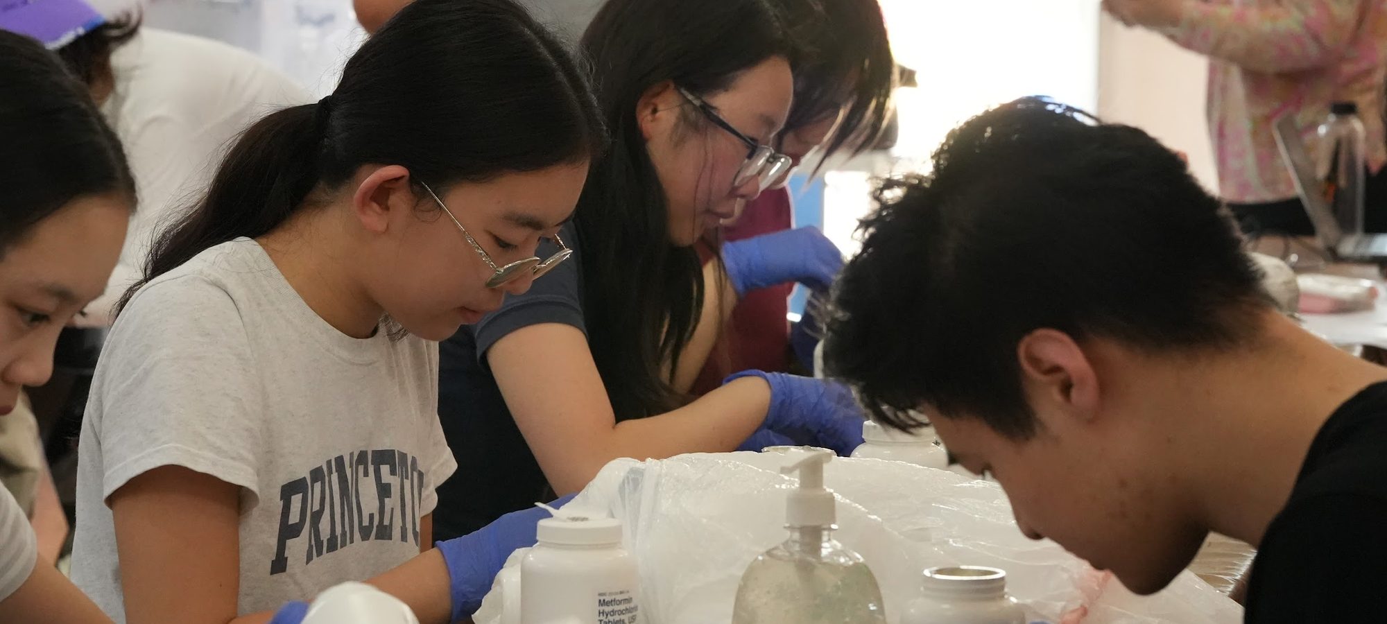 Teenage ACO Youth volunteers in gloves working shoulder to shoulder at the pharmacy table, counting and bagging medication